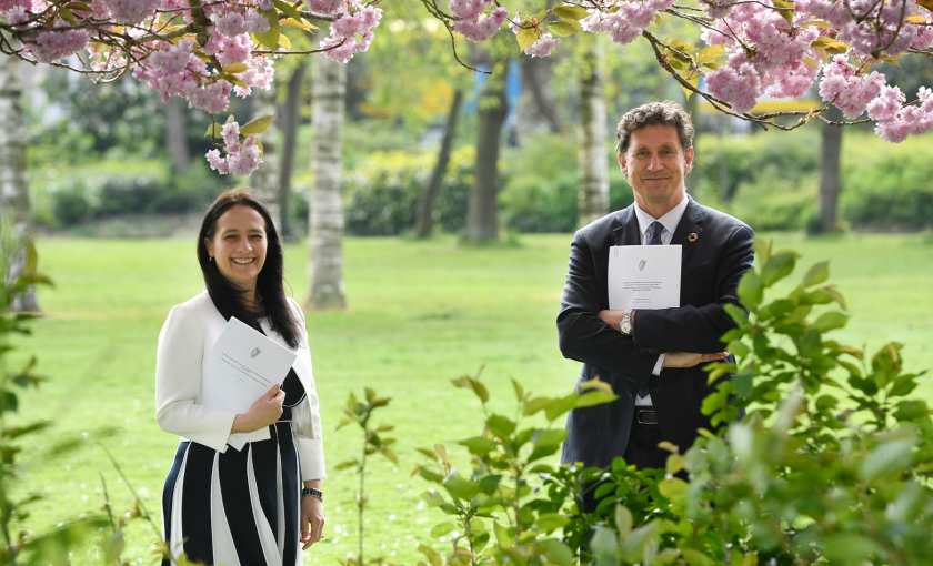A photo of two people standing in a park, holding A4 paper documents.