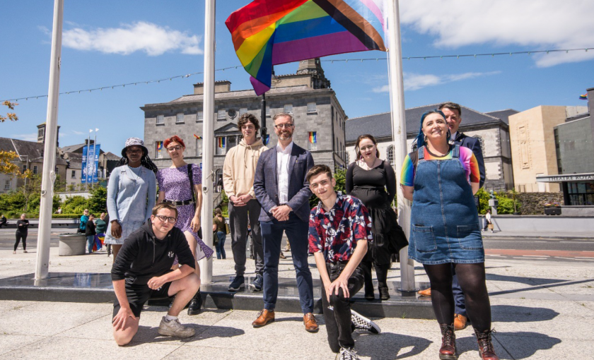 A photo of a group of people standing in front of a flag pole. The Pride rainbow flag flies from the pole. Roderic O'Gorman is standing in the centre of the group.