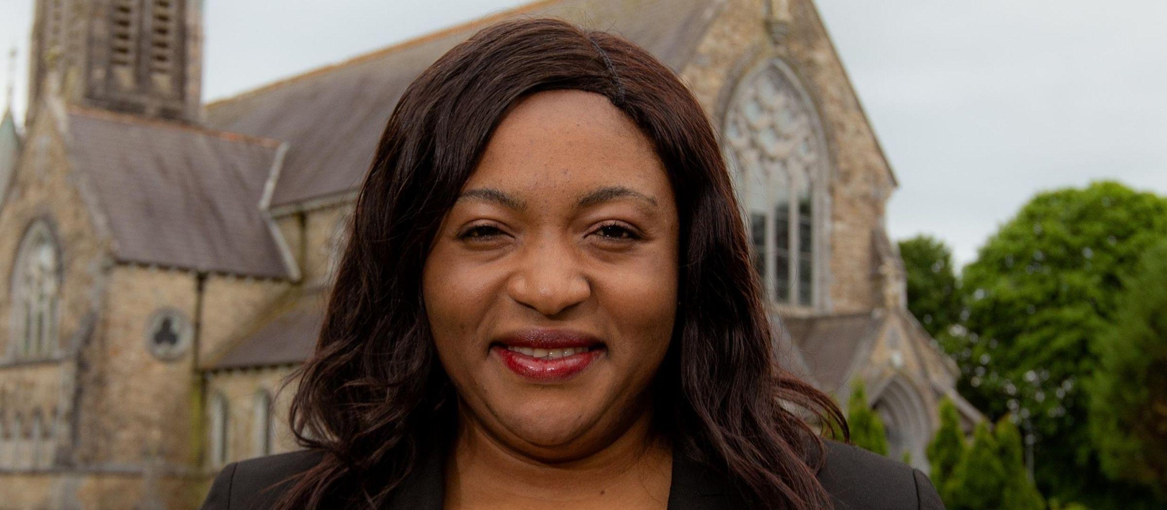 A woman with dark brown hair, Catherine Joseph, stands in front of a church.