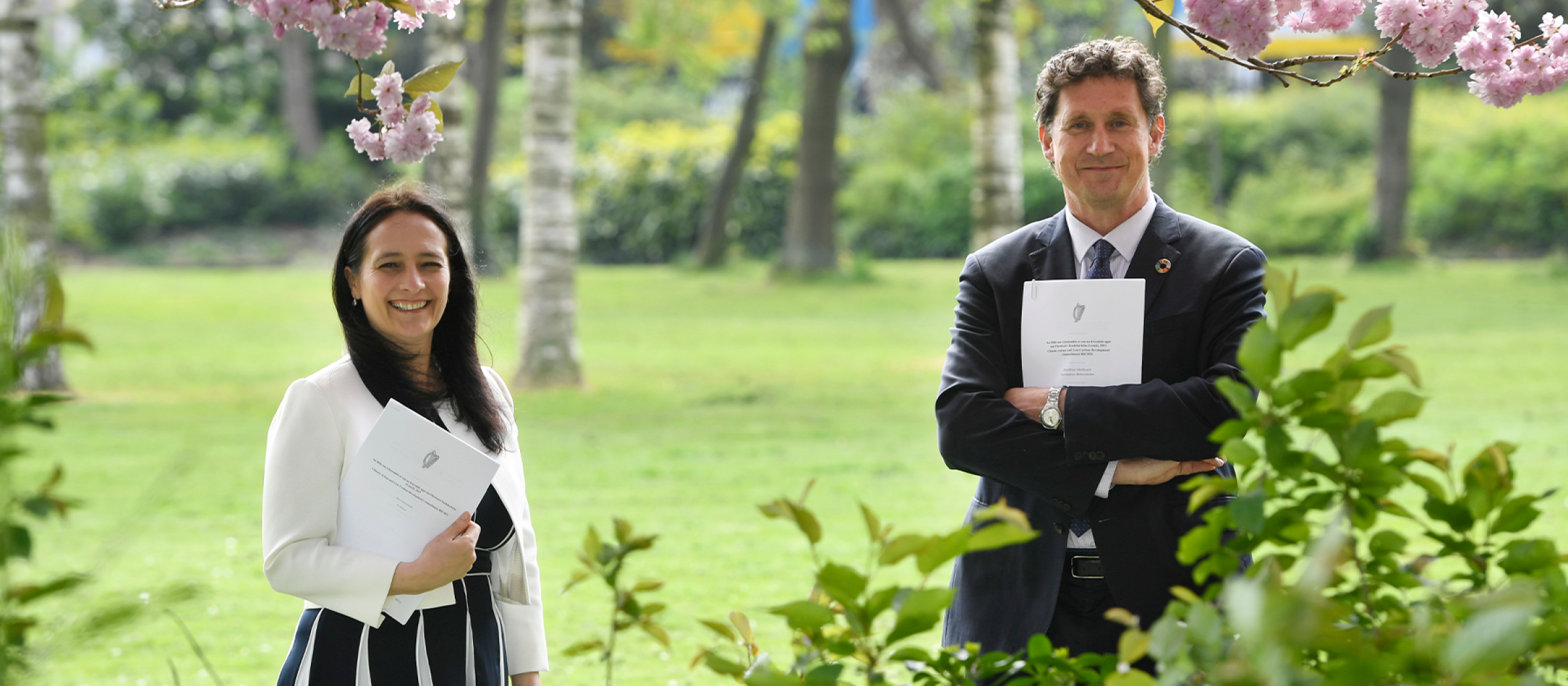 A photo of two people standing in a park, holding A4 paper documents.