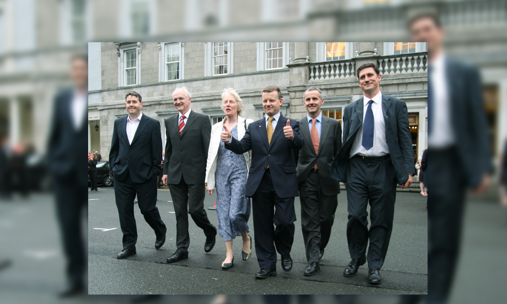 Six people in suits walk side by side on tarmac outside a stone building, Leinster House.