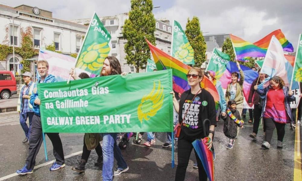 A group of people carrying Green Party and Rainbow Pride flags walk along a city street. 