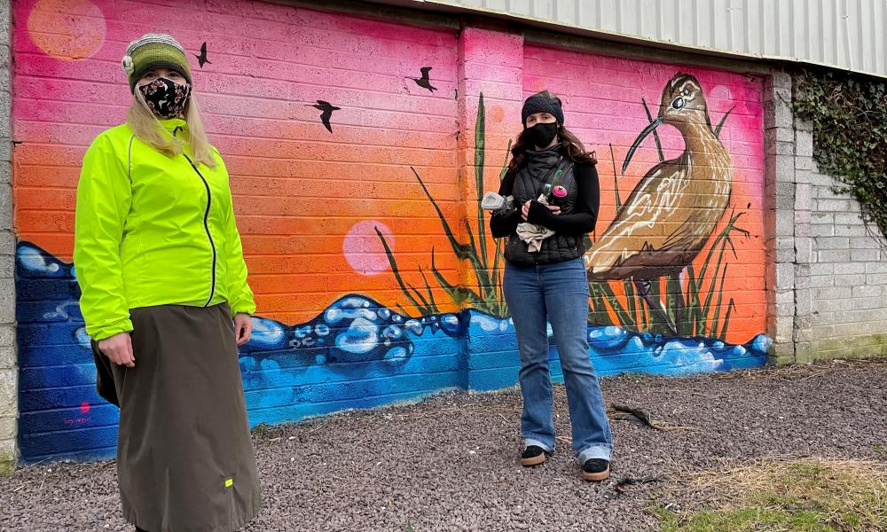 Two people wearing masks stand outside in front of a colourful mural of a curlew. There are oranges, pinks, blues, greens and browns.
