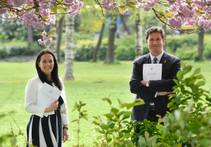 A photo of two people standing in a park, holding A4 paper documents.