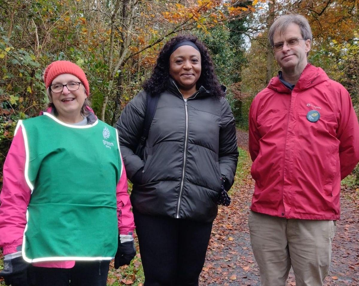 Three people, two women and a man, stand on a path in a woods. 